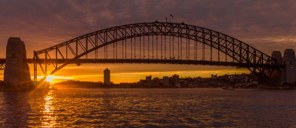 Sydney Harbour Bridge at sunset, with an orange sky and the sun going down to the left.